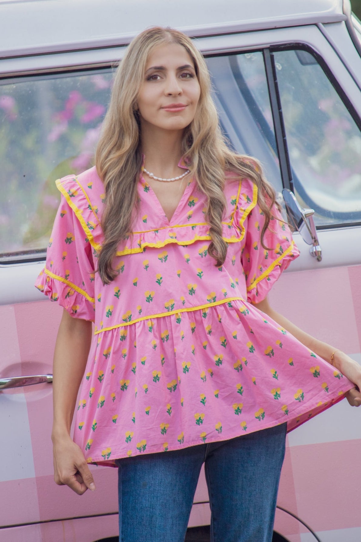 A woman wearing a pink tiered blouse with yellow and green floral prints and blue jeans, standing in front of a pink and white checkered van.