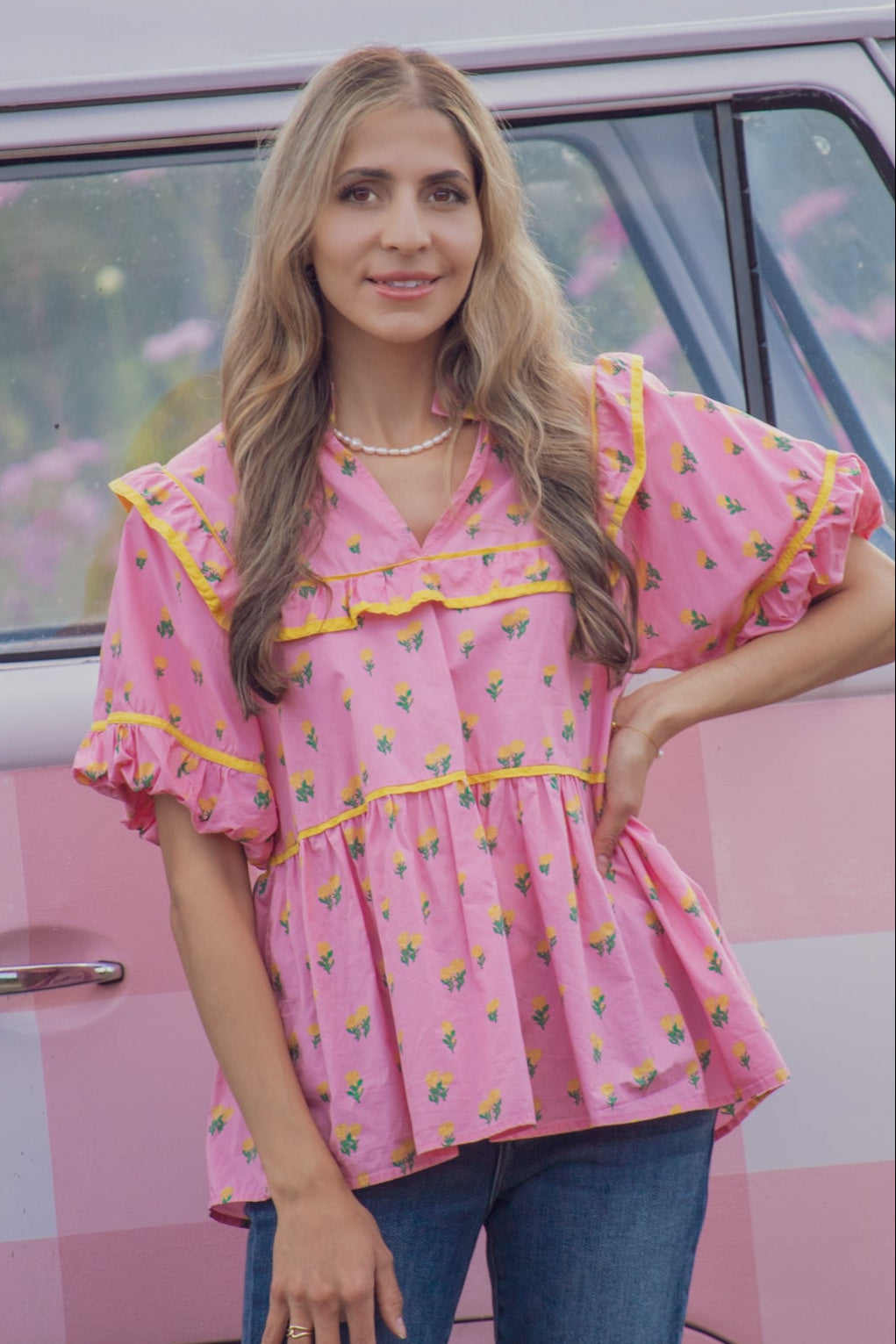 A woman wearing a pink short-sleeve peplum blouse with a small green and yellow floral print and yellow ruffle trim, paired with blue jeans, standing in front of a pink and white checkered van.